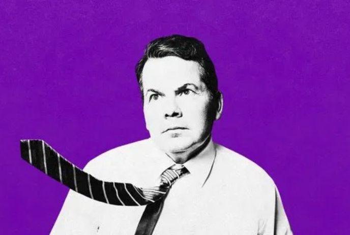 A black and white photo of Bruce McCulloch wearing a tie being blow upwards, behind a purple background.