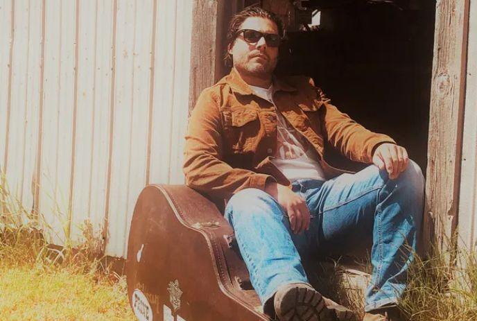 Ryan Naismith leaning on his guitar case outside on a Sunny day.