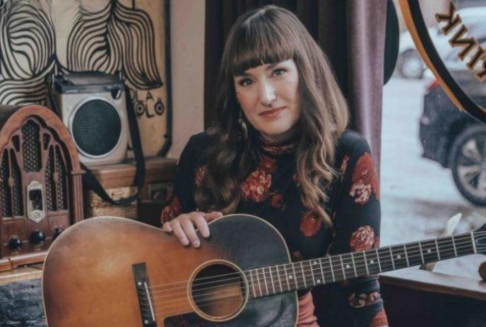 Suzie Ungerleider sitting down in a room, holding a guitar and looking at the camera.