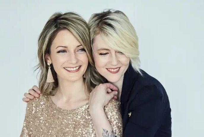 Amanda Walther and Sheila Carabine of the band Dala smiling and hugging each other, in front of a light blue background.