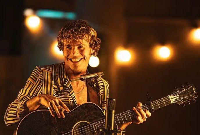 Kim Churchill performing on stage with a harmonica and guitar, in front of a light string.
