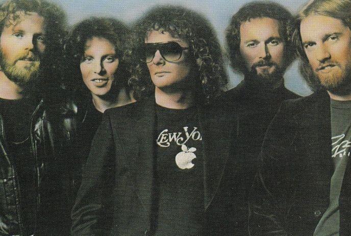 A vintage photo of the 5 band members of April Wine, wearing dark clothes and the middle one wearing sunglasses.