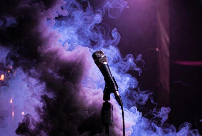 A microphone stands enveloped in swirling purple and blue smoke on a dimly lit stage.
