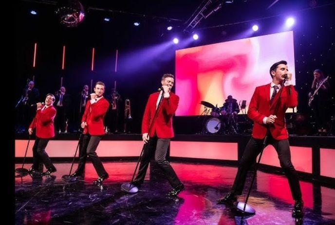 A group of male performers in red suits sing energetically on a vibrant stage.