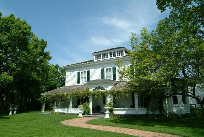 Eldon House house with green shutters and a wraparound porch. It is surrounded by lush trees and a well-manicured lawn.
