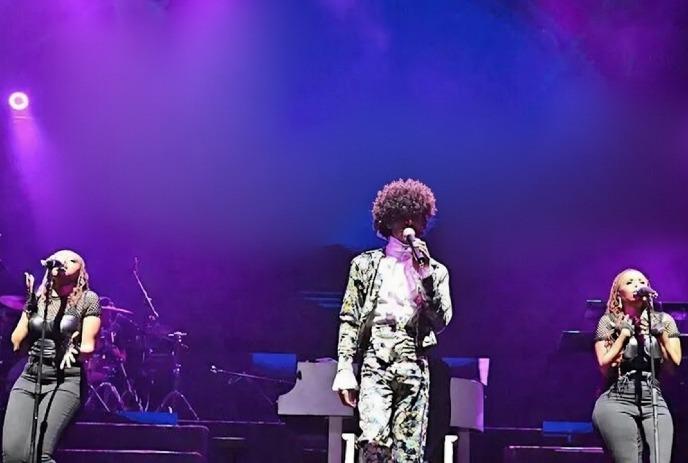 Three performers sing under vibrant purple stage lights.