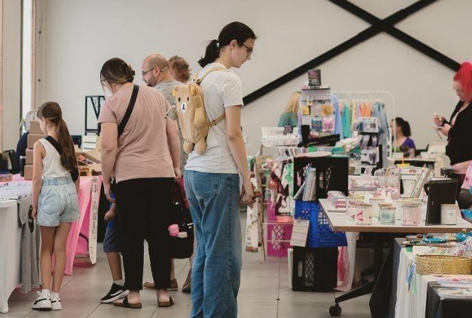 Indoor market scene with people browsing vendor tables of crafts, clothes, candles; cozy, lively atmosphere.