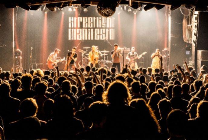 A packed crowd faces a band onstage, bathed in warm spotlights as hands rise and music fills the venue.