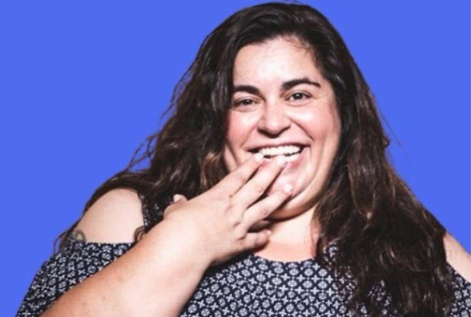 Debra is laughing with fingertips lightly touching chin and lips, smiling brightly before a solid blue background.