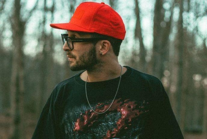 A side-profile portrait of Chris Webby rapper, standing outdoors with blurred woodland behind.