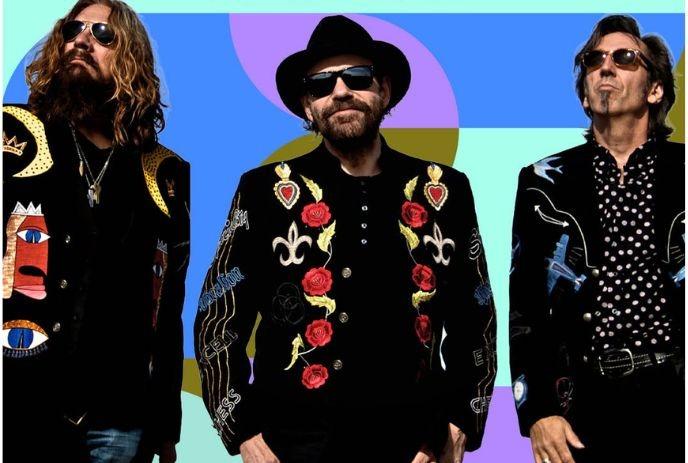 Three musicians in embroidered black jackets and sunglasses pose confidently against a bold, colorful geometric backdrop.