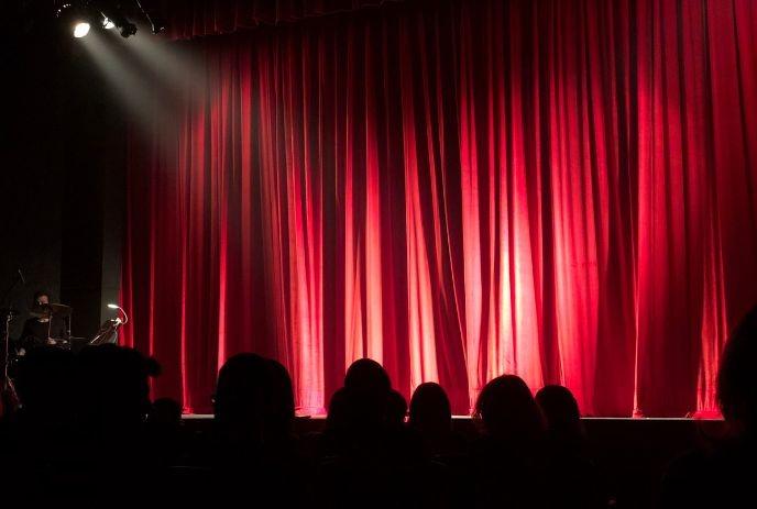 Red theatre curtains glow under bright spotlights as the audience waits.