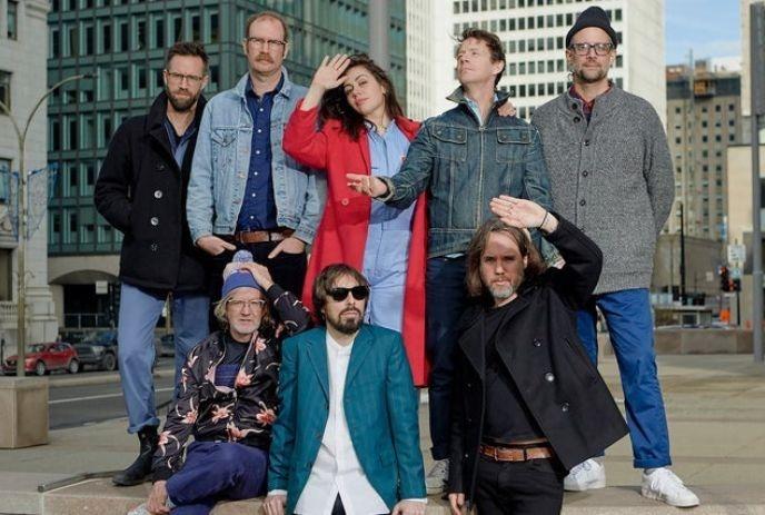 The 8 band members of Broken Social Scene, outside on a sunny day, posing in front of buildings in a city.