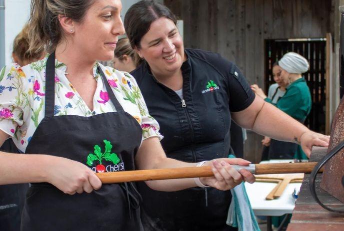 Two women in Growing Chefs uniforms placing pizzas in a wood-fire oven, with people in the background.