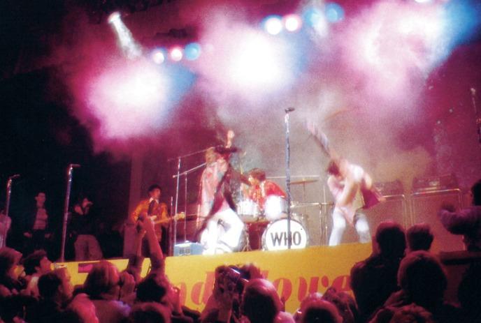 The Who jamming onstage with lights behind and a crowd in front of them.
