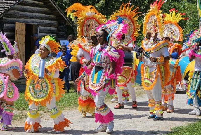 A group of people in vibrant, feathered costumes with elaborate headdresses parade outdoors near a log cabin on a sunny day.