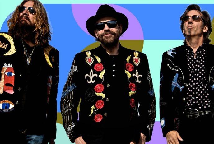 Three musicians in embroidered black jackets and sunglasses pose confidently against a bold, colorful geometric backdrop.