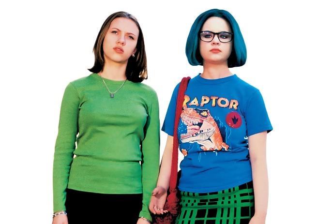 Two people stand together, one wearing a green top and black pants and one wearing a blue t-shirt with a green and black skirt.