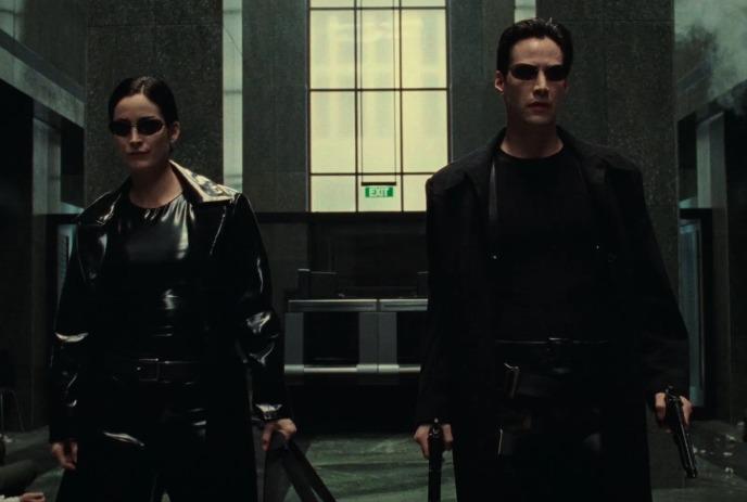 A man and a woman dressed in black holding gun in a dim hallway with a window in the background.