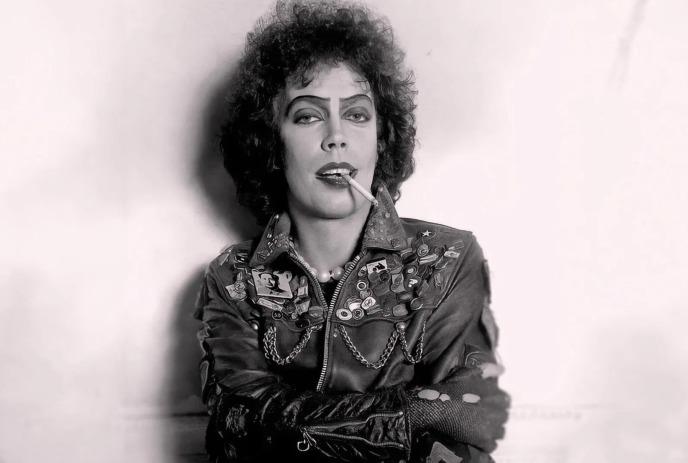 A person in a studded leather jacket with dramatic makeup poses confidently while holding a cigarette.