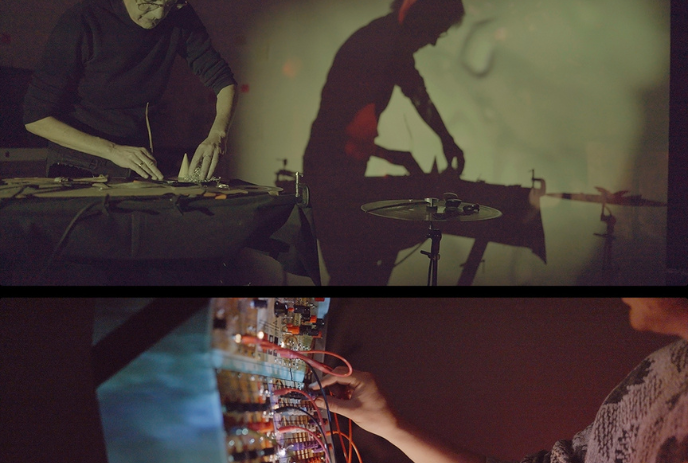 Two artists perform with experimental electronic instruments and visuals in a dimly lit setting.