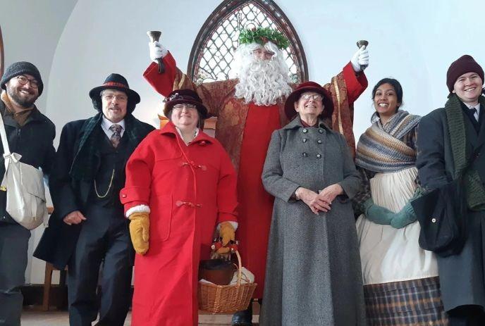 A cheerful group in 19th century-style coats stands in a church with St. Nick at the center.