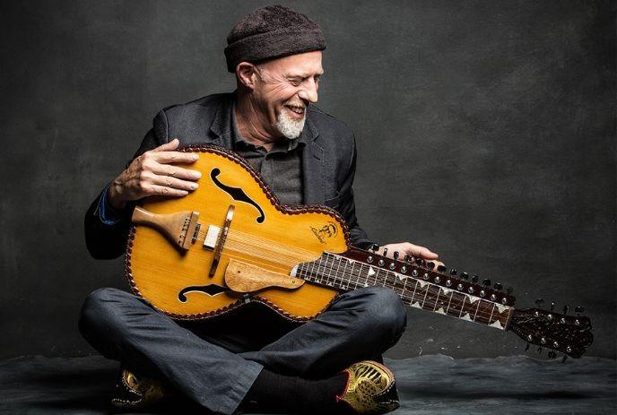 Harry Manx seating on the floor, holding a string instrument, looking down with a happy face expression.