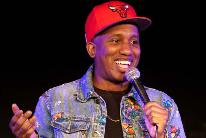 Chris Redd in red cap and floral denim jacket holding a microphone under a black background.