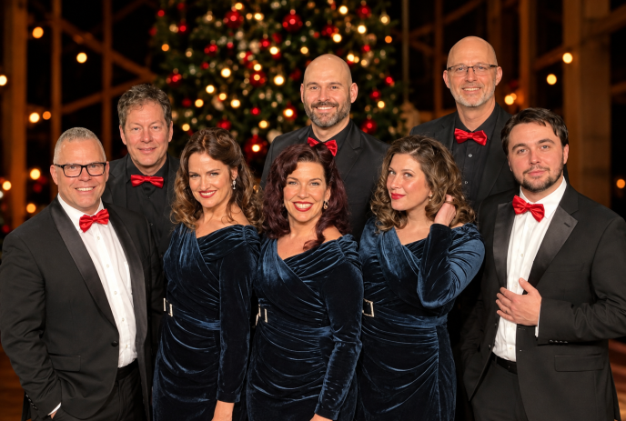 Eight performers in formal attire pose before a decorated Christmas tree with festive lights.