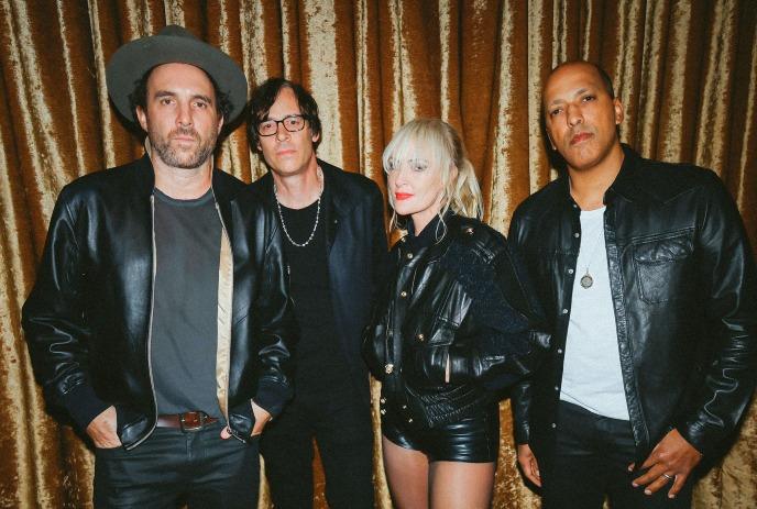 Four people stand side by side against a brown curtain backdrop. They wear black leather jackets, with neutral expressions.