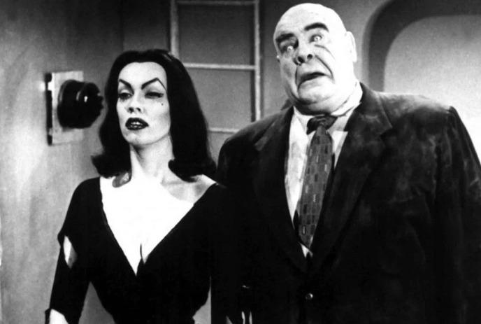 Black-and-white scene of a gothic woman and a wide-eyed, imposing man, evoking classic horror-comedy vibes.