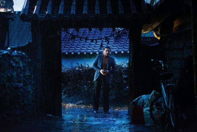 Man in suit holding camera stands in rain-soaked courtyard at night, lit by blue tones.