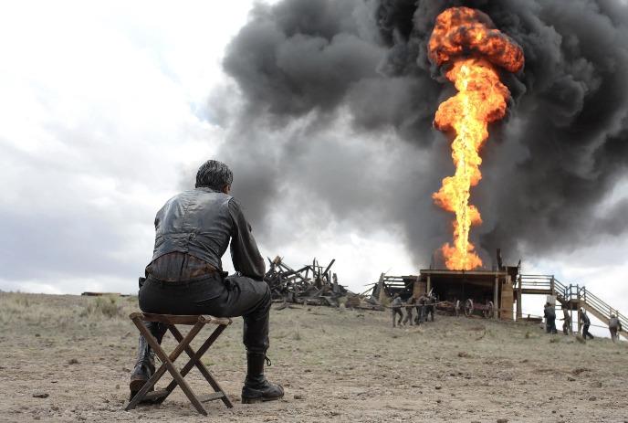 A man sits on a stool, watching a massive oil explosion billow fire and dark smoke in an open, desolate field.