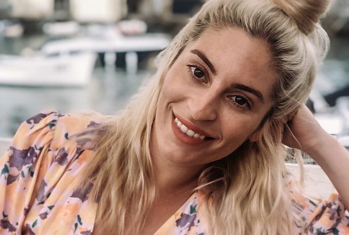 Jackie Pirico with light hair in a floral top, seated by a marina, leaning on one arm near boats.