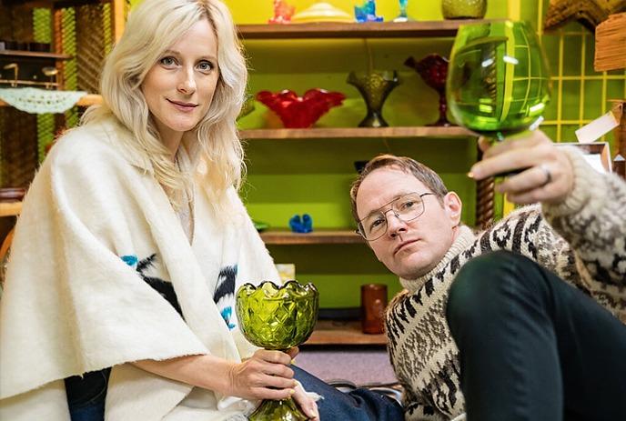 Two artists pose inside an ornate frame, each holding a green glass goblet in a colorful room.