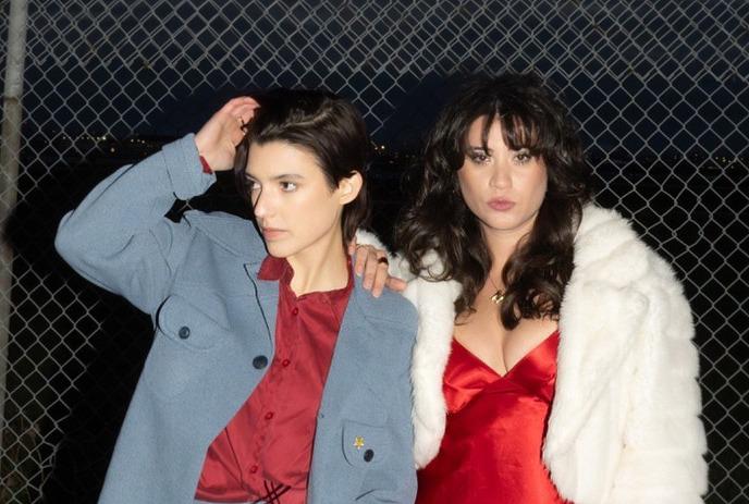 Two stylish people stand together at night, one in a blue suit and the other in red and white, behind a wire fence.