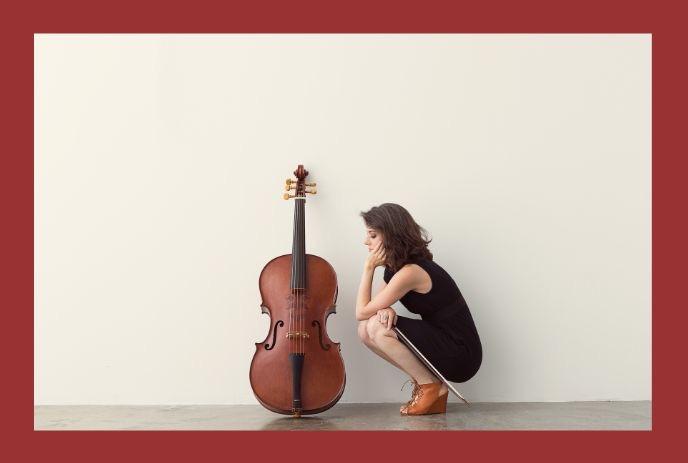 Elinor Frey, a cellist, kneels beside her cello against a plain wall with a red border.