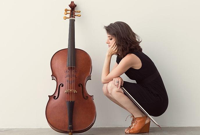 Elinor Frey, a cellist, kneels beside her cello against a plain wall