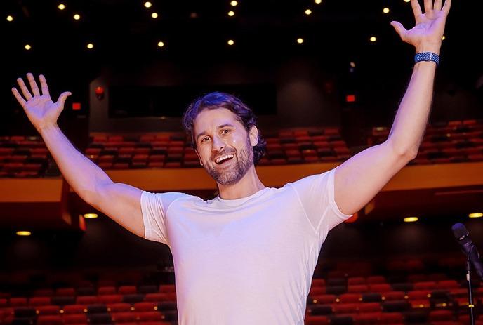Alex Mackenzie smiling with his arms in the air, on stage with empty theatre seats in the background.