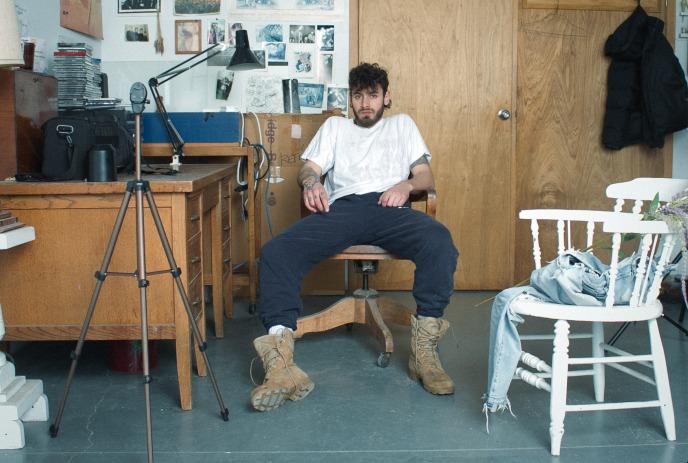 Allan Rayman sits on a wooden chair in a cluttered room with a desk, tripod, and wall covered in photos and art.