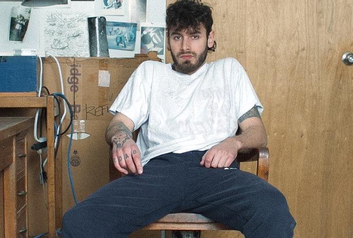 Allan Rayman sits on a wooden chair in a cluttered room with a desk, tripod, and wall covered in photos and art.