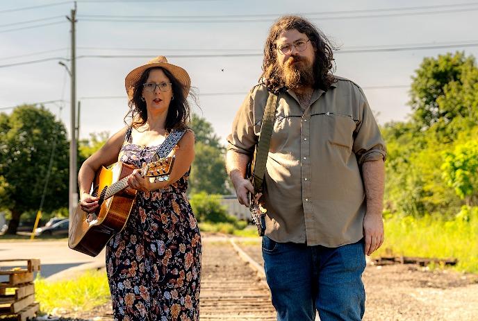 Two musicians walk along railway tracks, one carrying a guitar, with trees and greenery in the background.