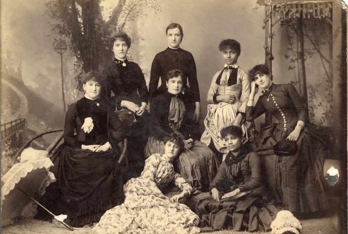 Vintage photo of eight people in Victorian-era clothing posing together in a garden setting.