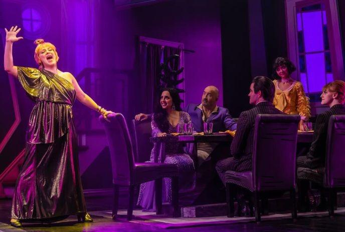 Six actors on stage; one stands in metallic gown; others seated at table under dramatic purple light.