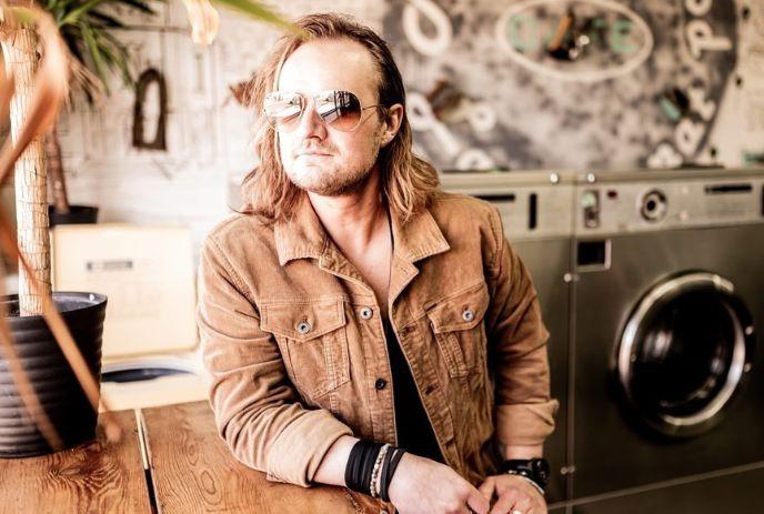 Artist in a tan suede jacket and sunglasses sits at table in laundromat, posing for a picture.