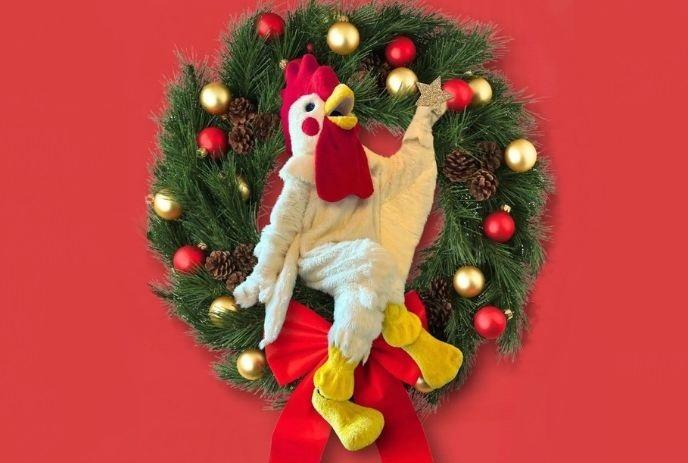 Holiday wreath with red bow and ornaments frames plush chicken figure under a red background.