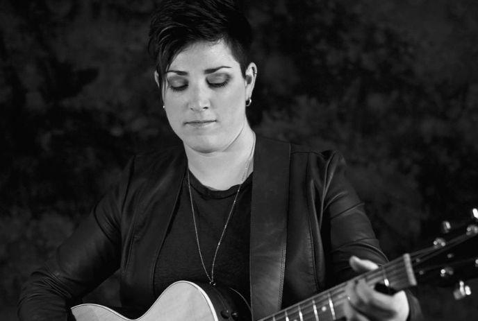 An artist playing an acoustic guitar in a black and white frame and a dark background.