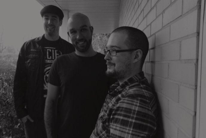 Three artists smiling and posing by a brick wall in a grayscale outdoor scene.
