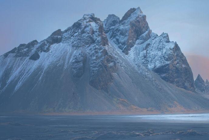 Snow-dusted jagged peaks rise at dusk; soft pink-blue sky glows above stark frozen plain.