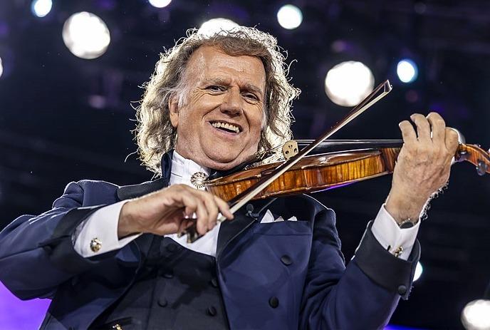 Violinist in tuxedo performing on stage before orchestra and glowing audience lights in concert hall ambience shines.